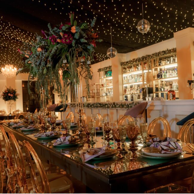 Mesa imperial con mobiliario elegante, centros de mesa y barra iluminada en el salón de Amara Home, hacienda para bodas y eventos